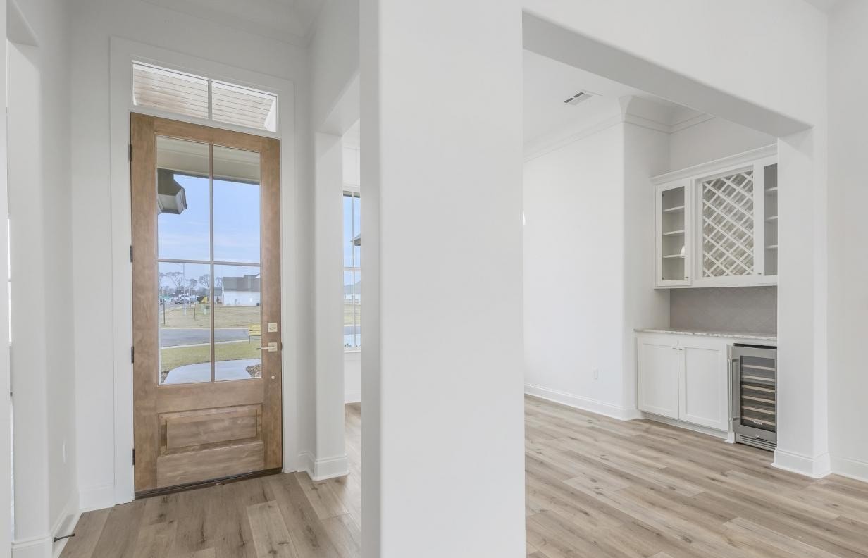 A modern entryway with a wooden front door, large glass panels, and light wood flooring; a partial view of a built-in dining area with a wine rack and wine fridge is visible to the right.