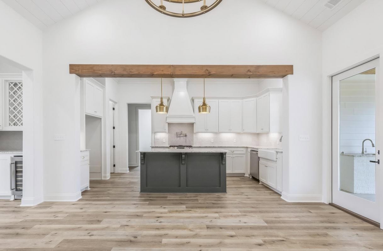 Modern kitchen with white cabinets, a dark island, gold pendant lights, light wood floors, white walls, and a wooden ceiling beam. Glass door is on the right and a wine rack is visible on the left.