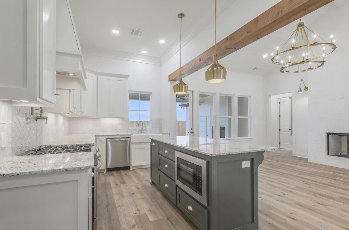 A modern kitchen with white cabinets, a gray island with a built-in microwave, stainless steel appliances, marble countertops, wood floors, pendant lights, a wooden ceiling beam, and an open view to the living area.
