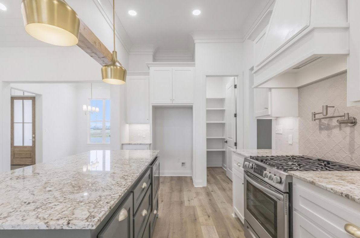 A modern kitchen with white cabinets, a marble island countertop, stainless steel appliances, gold pendant lights, a gas stove, a walk-in pantry, and light wood flooring.