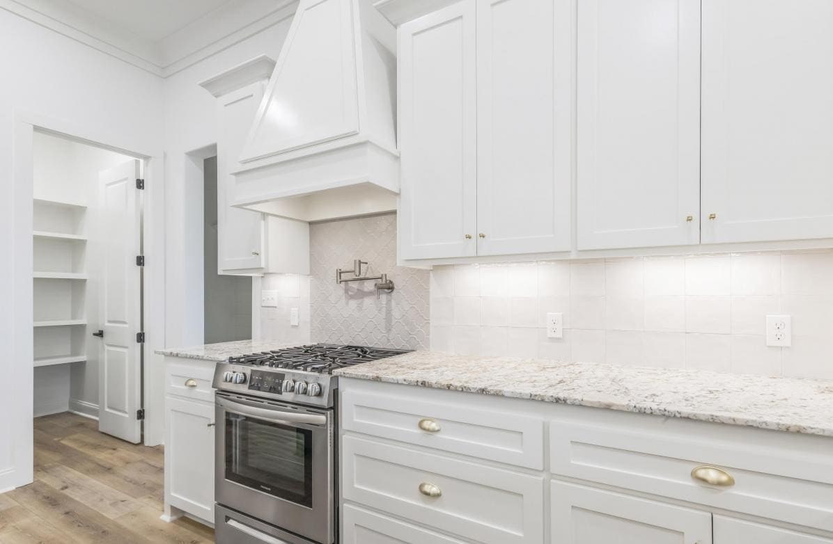 A modern kitchen with white cabinets, a stainless steel gas stove, a tile backsplash, white countertops, under-cabinet lighting, and a pantry visible through an open doorway.