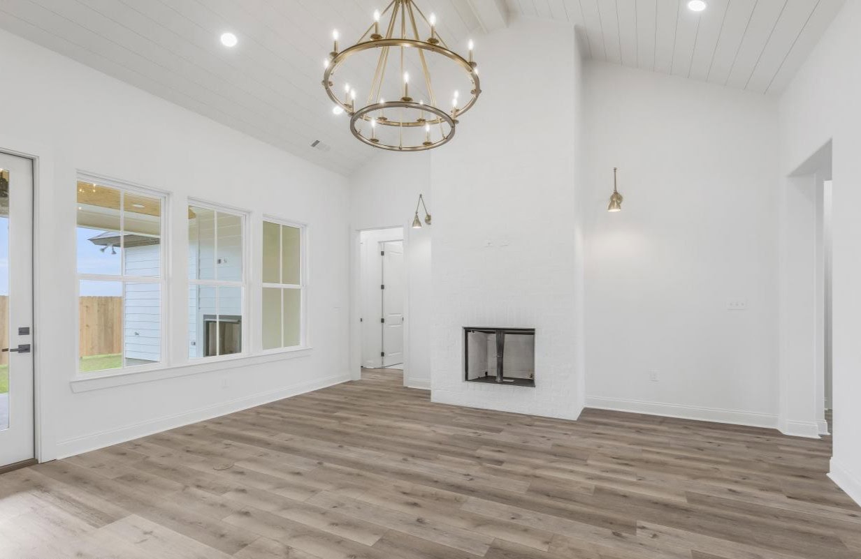 A bright, empty living room with white walls, a modern fireplace, wood-look flooring, large windows, a gold chandelier, and recessed ceiling lights. A door leads outside and there are wall sconces by the fireplace.