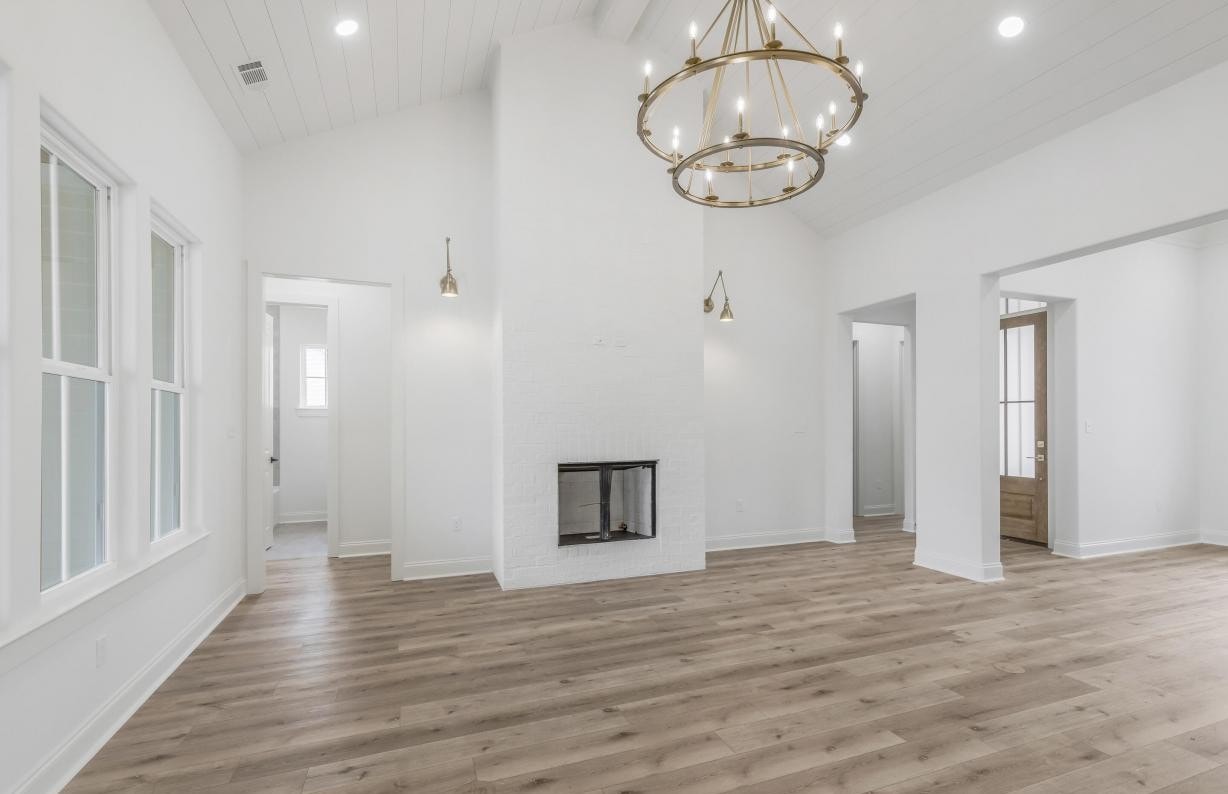 A spacious, empty living room with white walls, a white brick fireplace, wood flooring, high ceilings, a modern gold chandelier, and large windows letting in natural light.