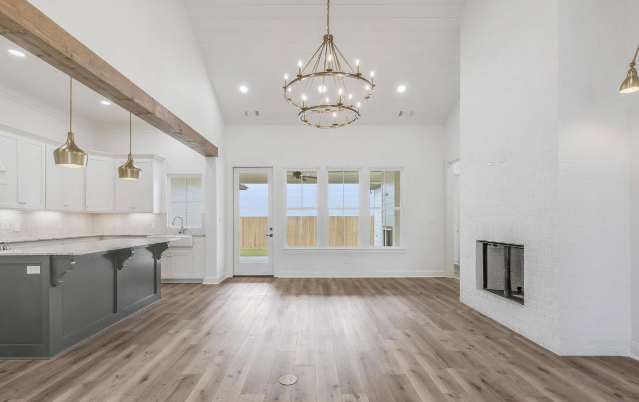 A spacious open-plan kitchen and living area with wood floors, white walls, pendant lights, a large chandelier, a white brick fireplace, and windows overlooking a fenced backyard.