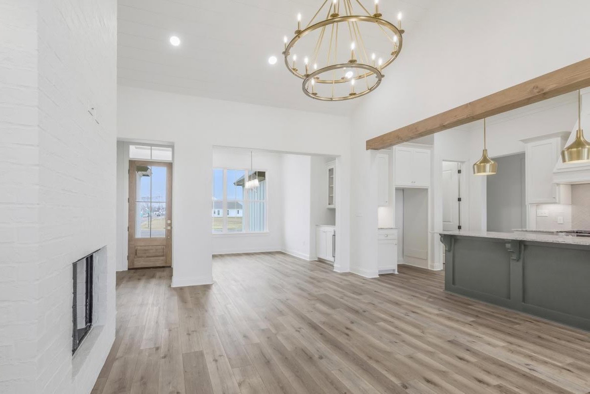 A spacious, empty modern living and dining area with light wood flooring, white walls, large windows, and gold light fixtures. The kitchen has an island with a gray base and white cabinetry.