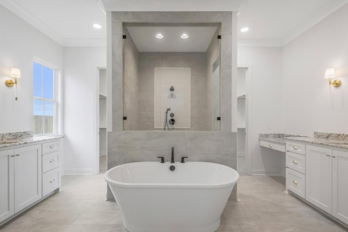 Modern bathroom with a freestanding white bathtub in the center, black fixtures, a walk-in shower behind the tub, double sinks on each side, white cabinetry, and wall-mounted lights. Natural light enters through a window.