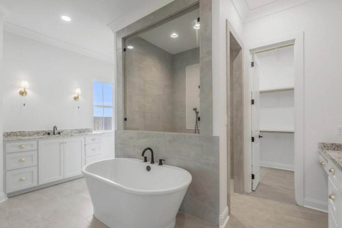 A modern bathroom with a freestanding white bathtub, a large glass-enclosed shower, double vanities with white cabinets and granite countertops, and a walk-in closet visible through an open doorway.