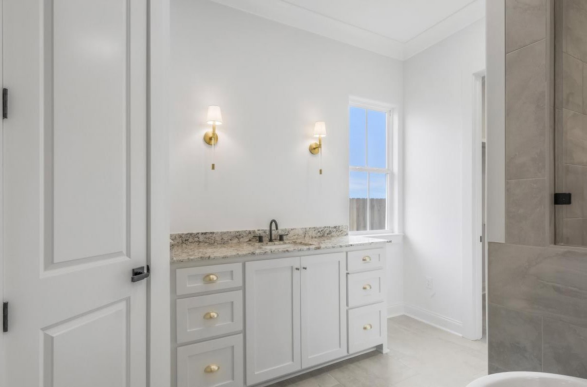 A bathroom with white walls, a white vanity with gold hardware, a granite countertop, two wall-mounted light fixtures, a window, and a partial view of a gray tiled shower.