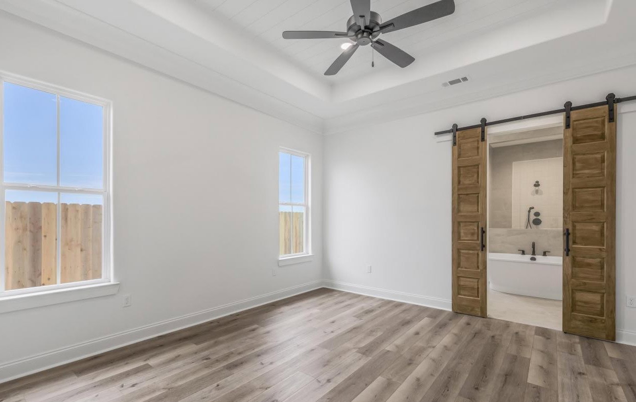 A bright, empty room with light wood flooring, white walls, two tall windows, a ceiling fan, and wooden sliding barn doors leading to a bathroom with a bathtub.