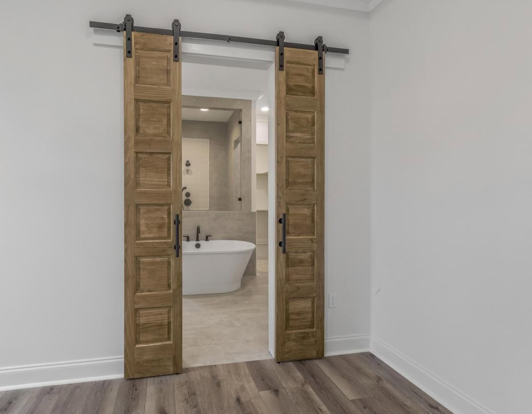A room with light wood flooring and white walls features double sliding wooden barn doors that are open, revealing a bathroom with a freestanding white bathtub and tiled floor and walls.