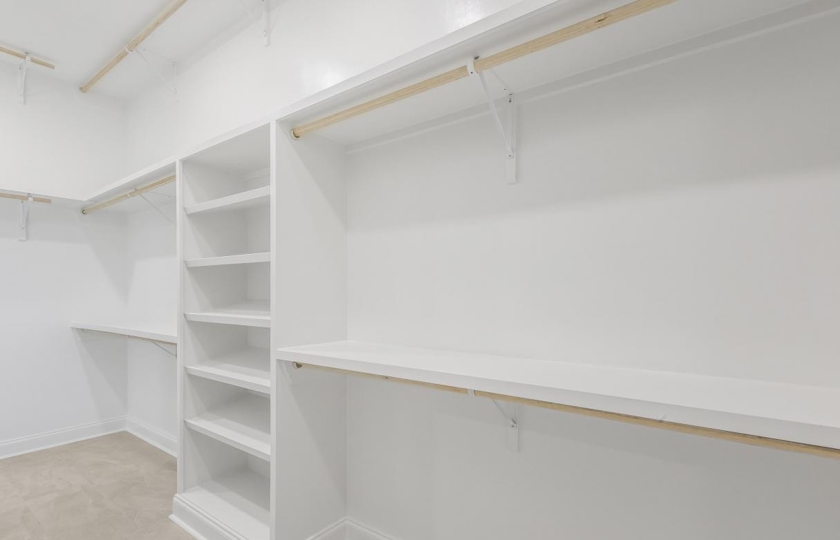 Empty white walk-in closet with built-in shelves, open hanging rods, and beige carpet flooring; walls, shelving, and ceiling are all white.