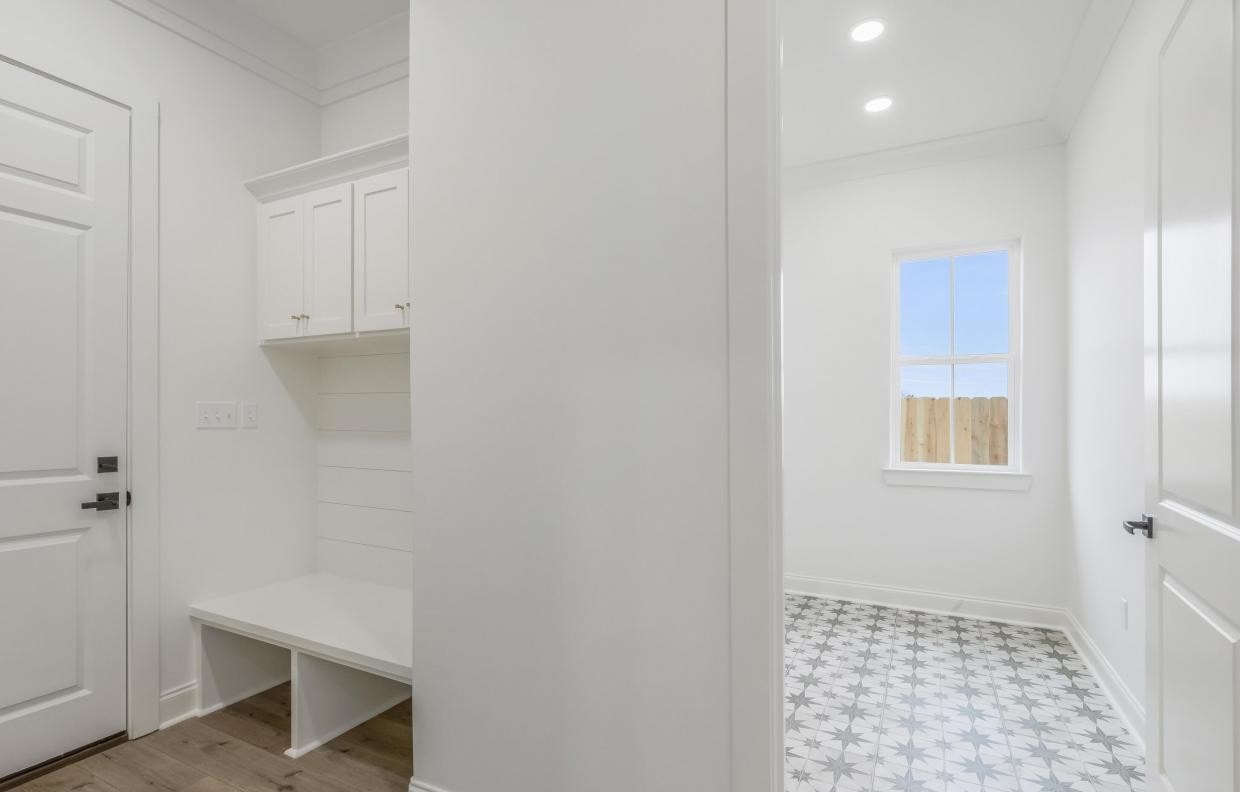 A white mudroom with built-in cabinets and bench is adjacent to a bright laundry room with patterned tile flooring, white walls, a window, and a wooden fence visible outside.