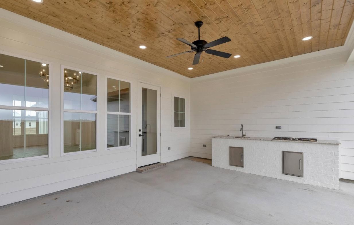 A covered patio with a wood-paneled ceiling, ceiling fan, white horizontal siding, large windows, a glass door, and an outdoor kitchen area with a sink and built-in cabinets.