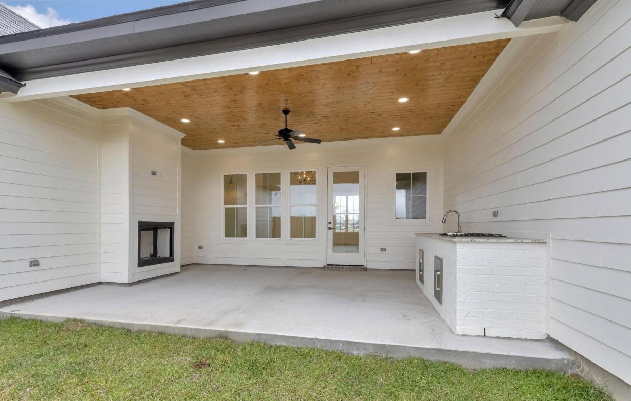 Covered patio with white siding, a wooden ceiling with recessed lights, a ceiling fan, a built-in outdoor fireplace on the left, and a small outdoor kitchen with a sink and grill on the right, overlooking a grassy yard.