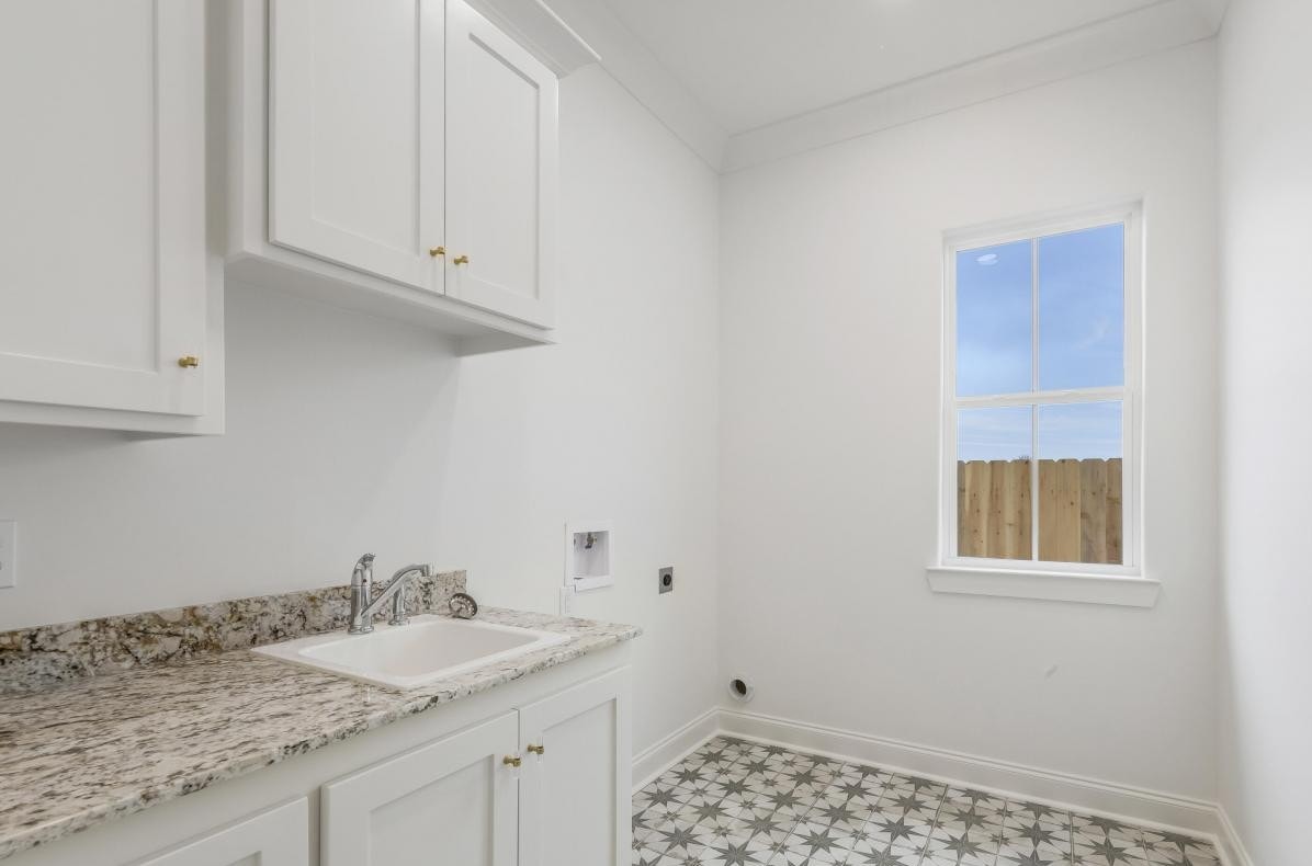 A laundry room with white cabinets, a granite countertop with a sink, patterned tile flooring, and a single window letting in natural light. There are outlets and hookups for a washer and dryer.