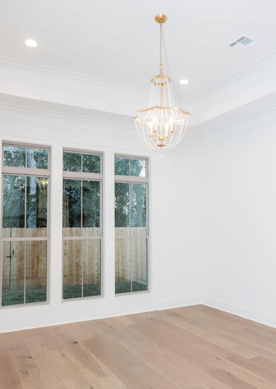 A bright, empty room with white walls, light wood flooring, three large windows showing a fenced yard, and a gold chandelier with hanging beads on the ceiling.