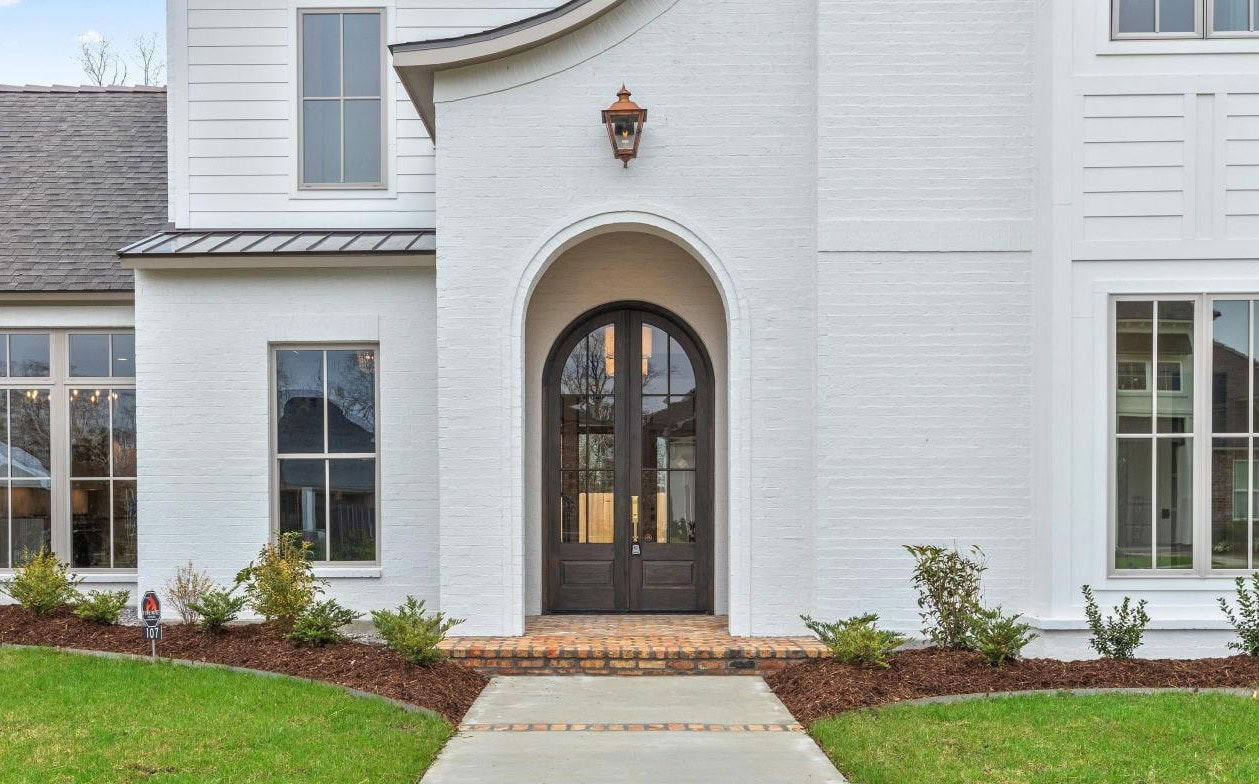 A modern white brick house with a central arched doorway, black framed double doors, large windows on each side, a lantern above the entrance, and a landscaped front yard with a concrete path and shrubs.