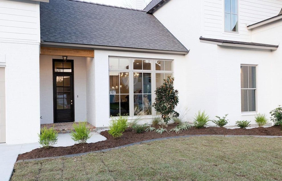 A modern white house with a large front window, dark front door, small porch area, landscaped garden bed with young plants, and a well-maintained lawn.