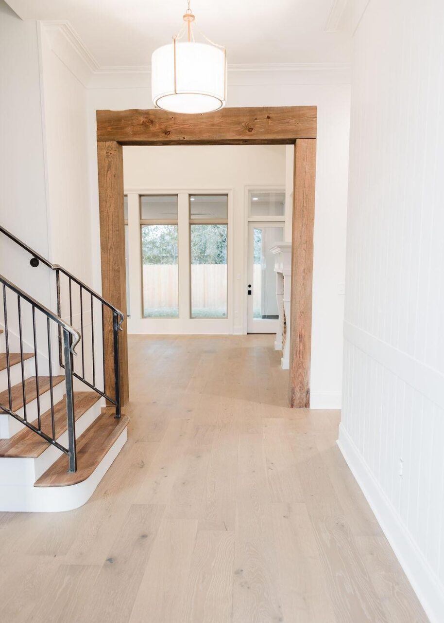 A modern, bright entryway with light wood floors, a staircase with wood steps and black railing on the left, a large wooden doorway frame, and floor-to-ceiling windows in the background.