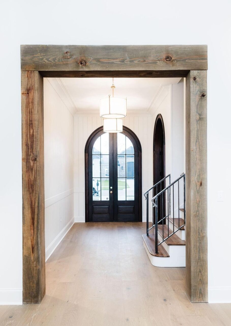 A bright entryway with light wood floors, rustic wood trim framing the doorway, and black double arched doors with glass panels. A small staircase with a metal railing is on the right, and a modern pendant light hangs above.