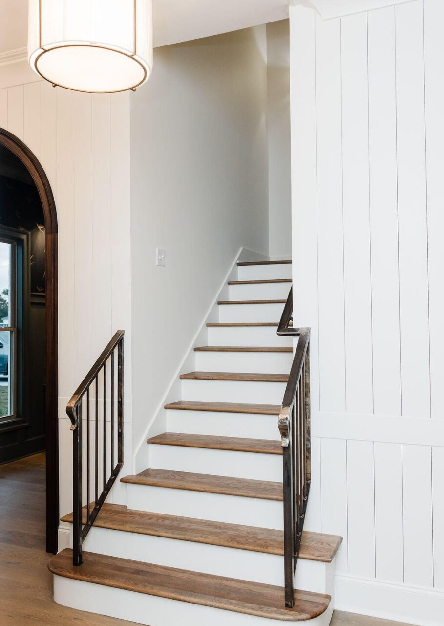 A staircase with wooden treads and white risers leads upward alongside a white paneled wall. Black metal railings are on both sides, and a large round light fixture hangs above. An arched doorway is visible to the left.