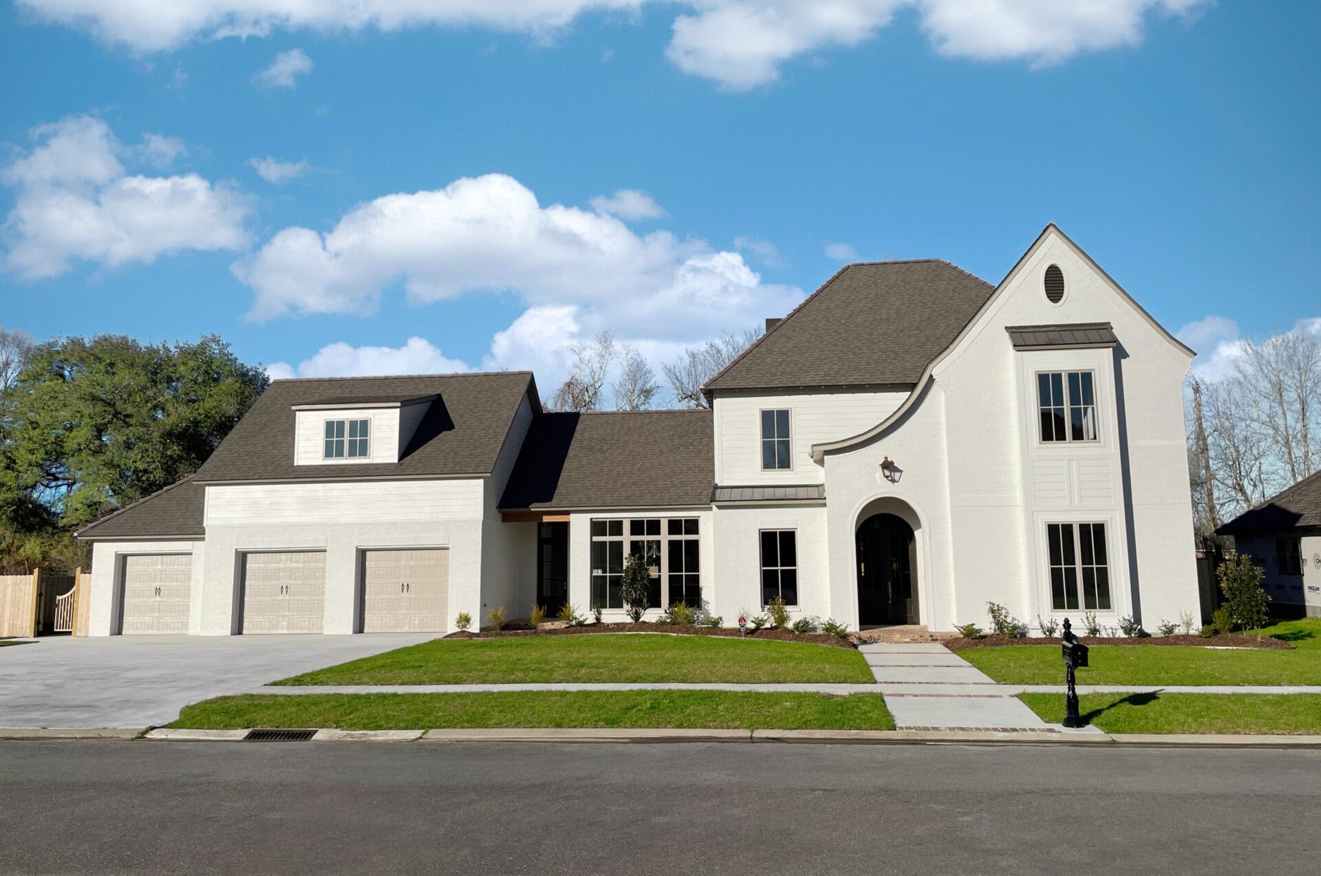A large two-story modern house with light-colored brick exterior, multiple tall windows, a three-car garage, manicured lawn, and a clear blue sky in the background.