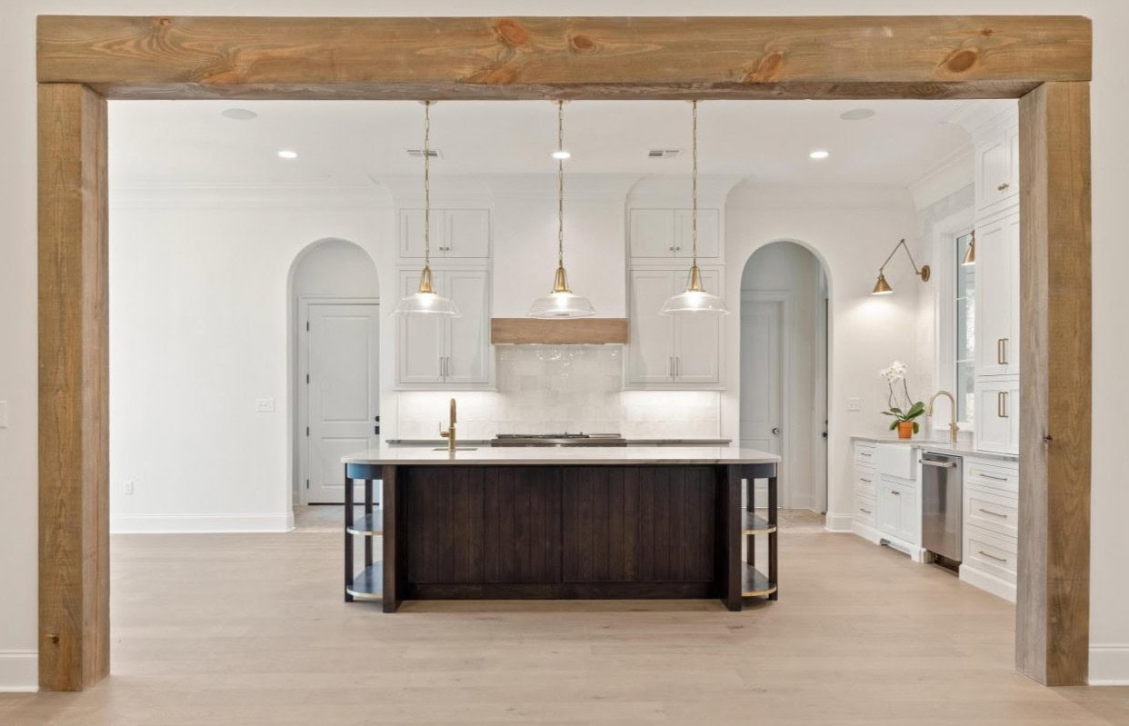 Modern kitchen with a large wooden frame, a dark central island with a white countertop, three pendant lights above, white cabinets, stainless steel appliances, and light wood flooring.