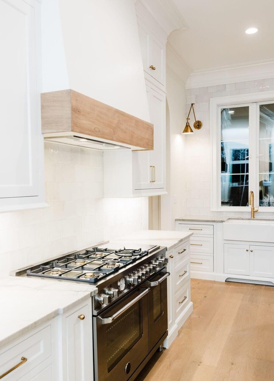 A modern kitchen with white cabinets, a stainless steel gas stove, light wood flooring, a white tile backsplash, a farmhouse sink, a brass faucet, and a gold wall sconce by a window.