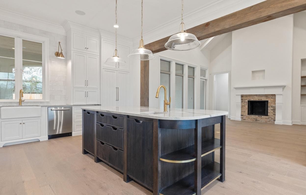 Modern kitchen with a dark wood island, gold faucet, white countertops, pendant lights, white cabinetry, built-in sink, stainless steel appliances, and an open living area with a fireplace in the background.