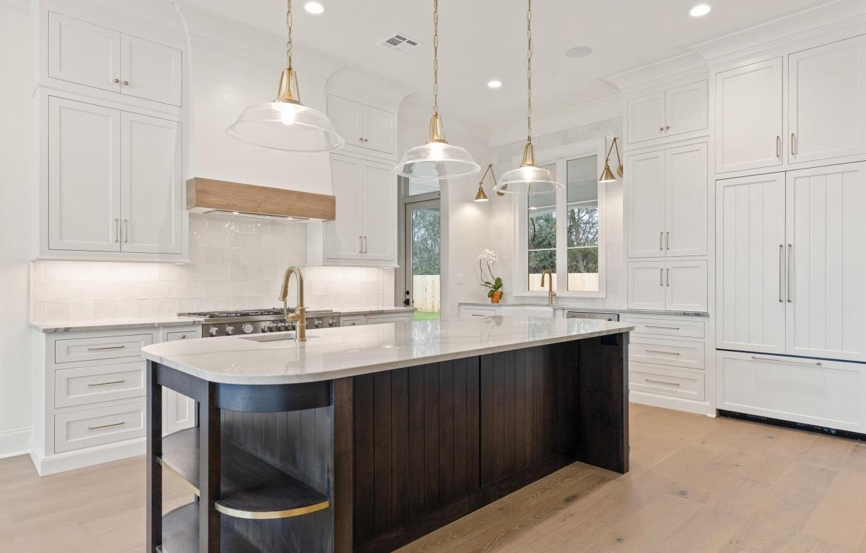 Modern kitchen with white cabinets, a large dark wood island, marble countertops, pendant lights, stainless steel appliances, and light wood flooring. A window provides natural light. Fruit bowl on the counter.