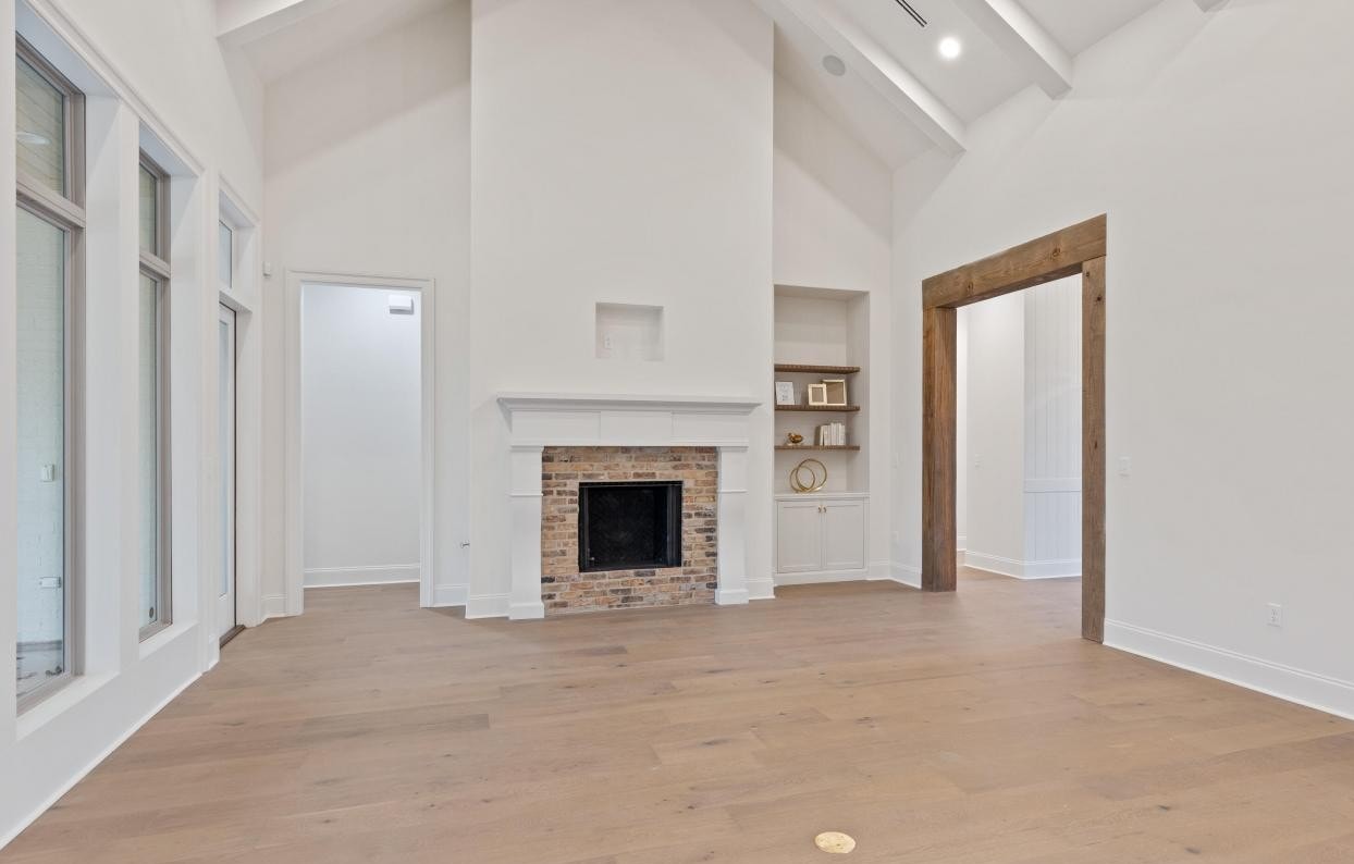 A spacious, empty living room with light wood flooring, a white brick fireplace, built-in shelves, high vaulted ceilings, and large windows. There is a wide doorway with a wooden frame leading to another area.