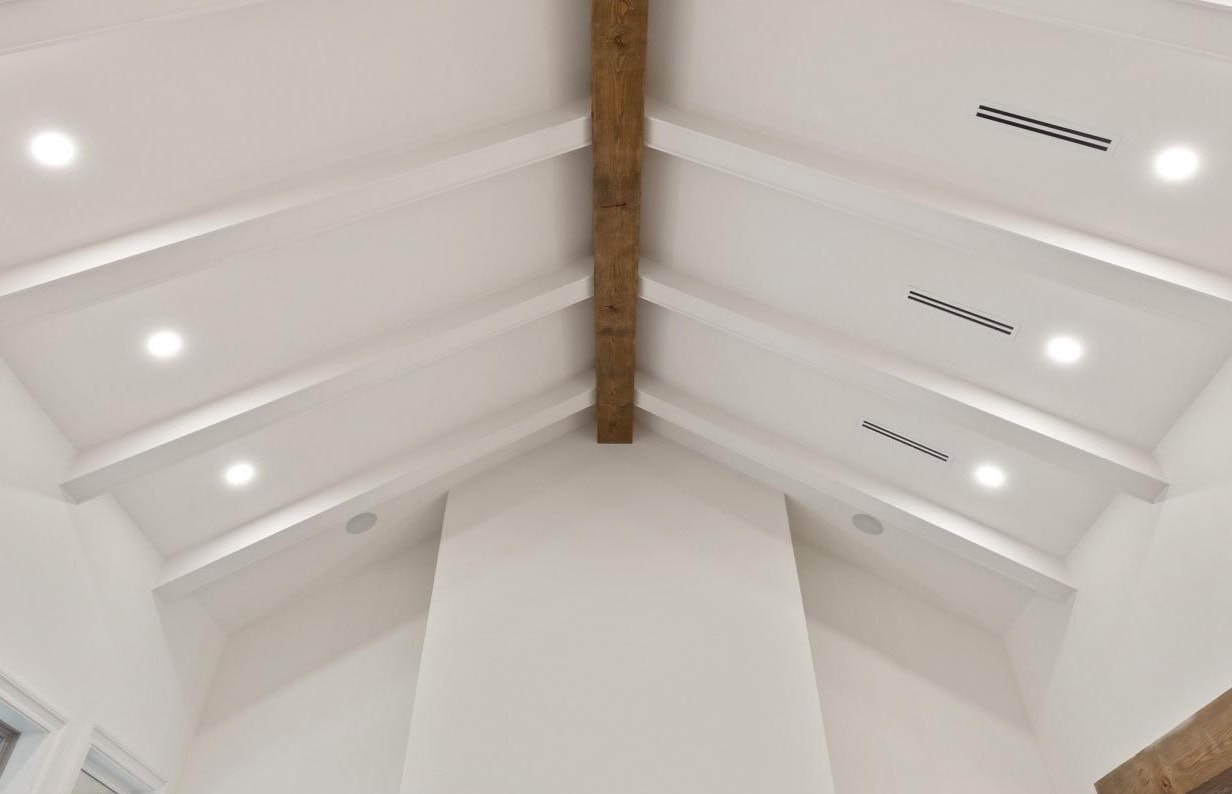 White vaulted ceiling with exposed wooden beam, recessed lighting, and linear air vents. The ceiling features symmetrical beams and a modern, minimalist design.