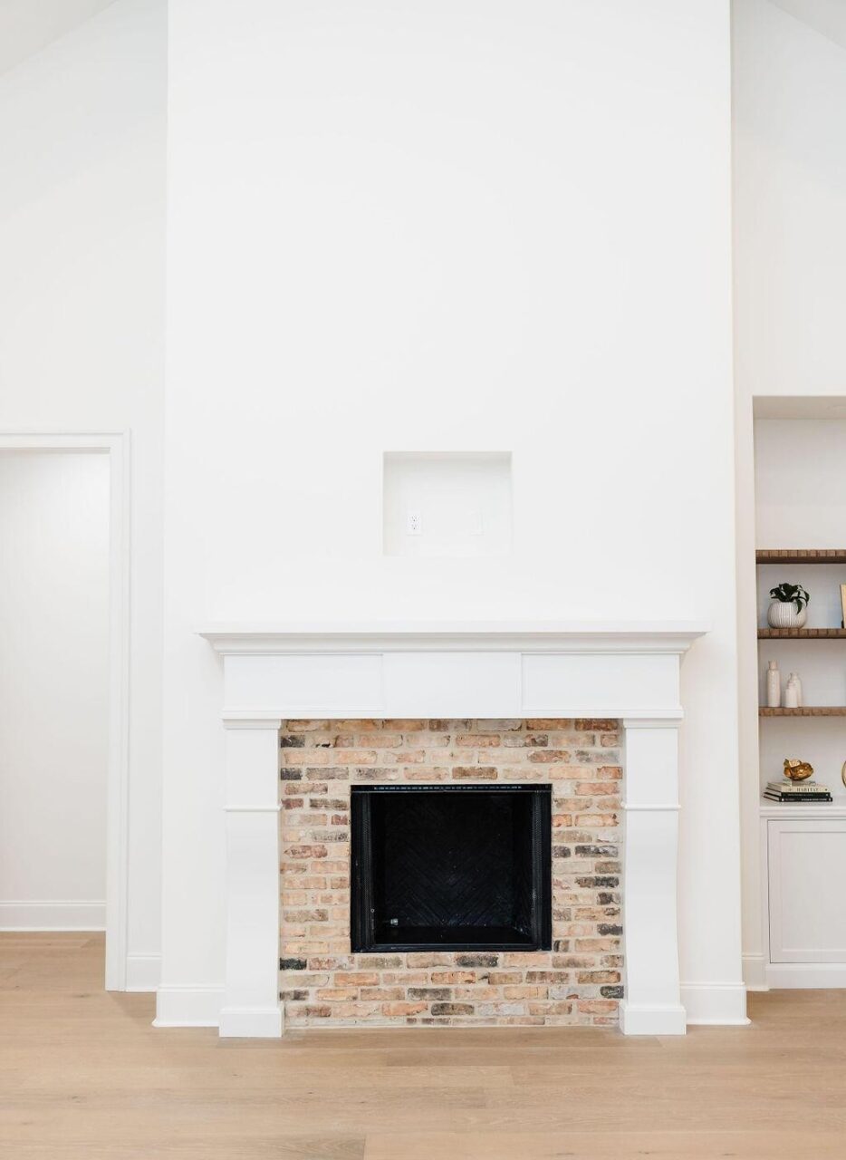A minimalist living room features a brick fireplace with a white mantel, flanked by built-in shelves and a doorway. The walls and trim are painted white, and the floor is light wood.