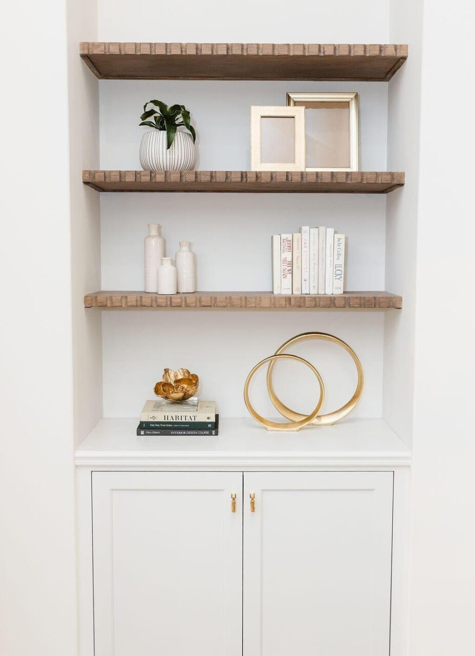 A built-in white shelving unit with three wooden shelves displays a potted plant, framed pictures, small vases, stacked books, and decorative gold sculptures above a white cabinet with gold handles.