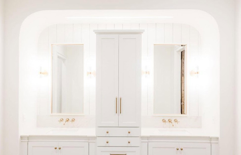 A bright, white bathroom features two sinks with gold faucets, two mirrors, and a tall central cabinet with gold handles. Vertical paneling and wall sconces are present on either side of the mirrors.