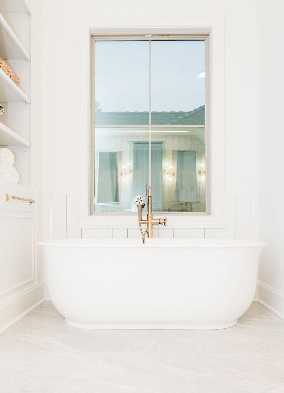 A white freestanding bathtub with a gold faucet is positioned below a large window. Shelves with towels and baskets are on the left, and the walls and floor are light-colored for a clean, modern look.