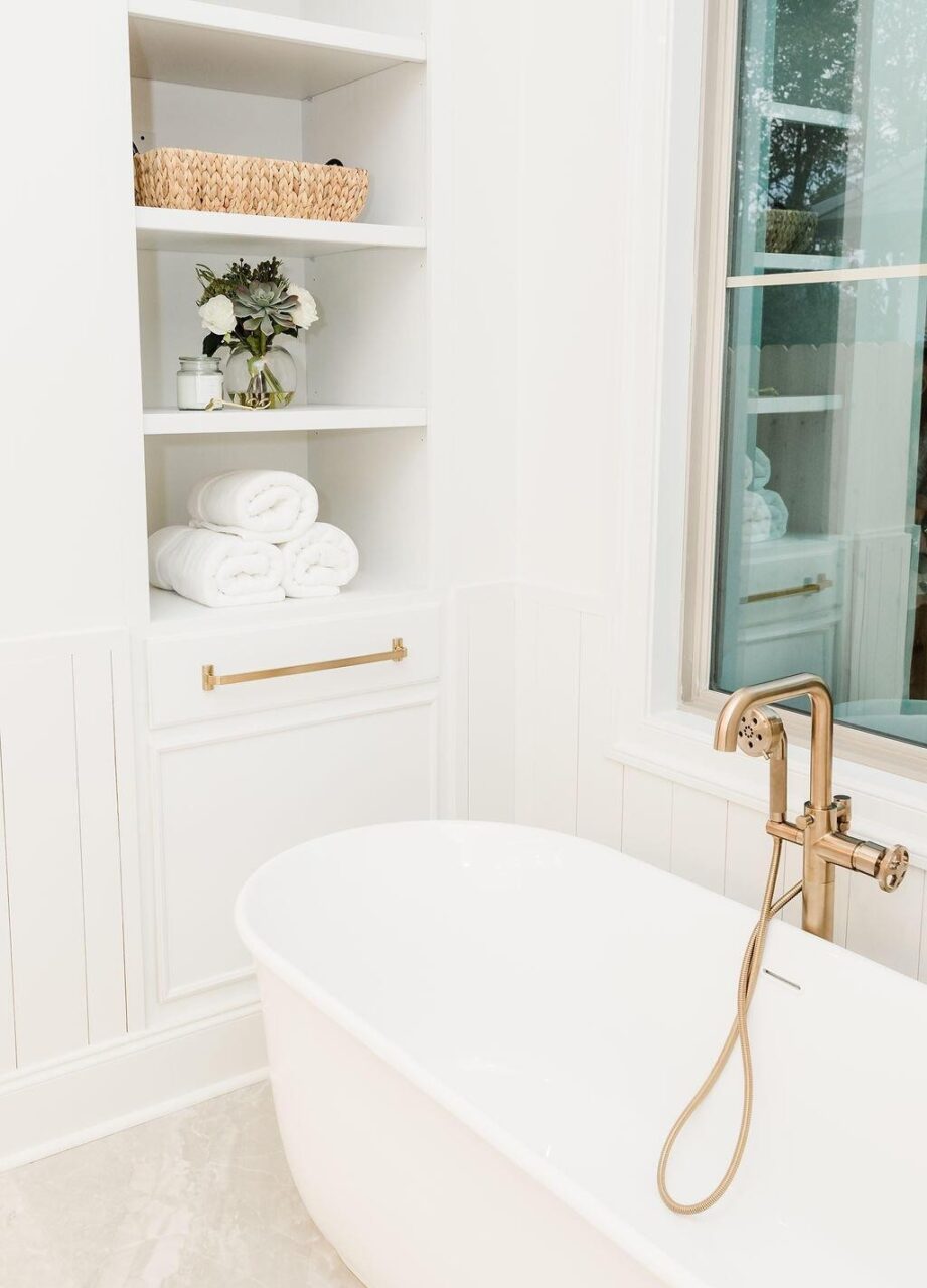 A white bathroom features a freestanding bathtub with a gold faucet, built-in shelves holding rolled white towels, a basket, and a small vase with flowers next to a large window.