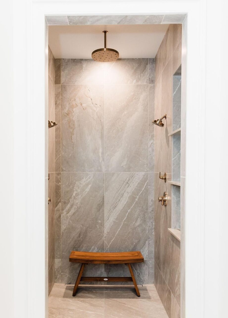 A walk-in shower with large gray marble tiles, a ceiling-mounted rainfall showerhead, wall-mounted shower controls, built-in shelves on the right, and a wooden bench placed on the floor.