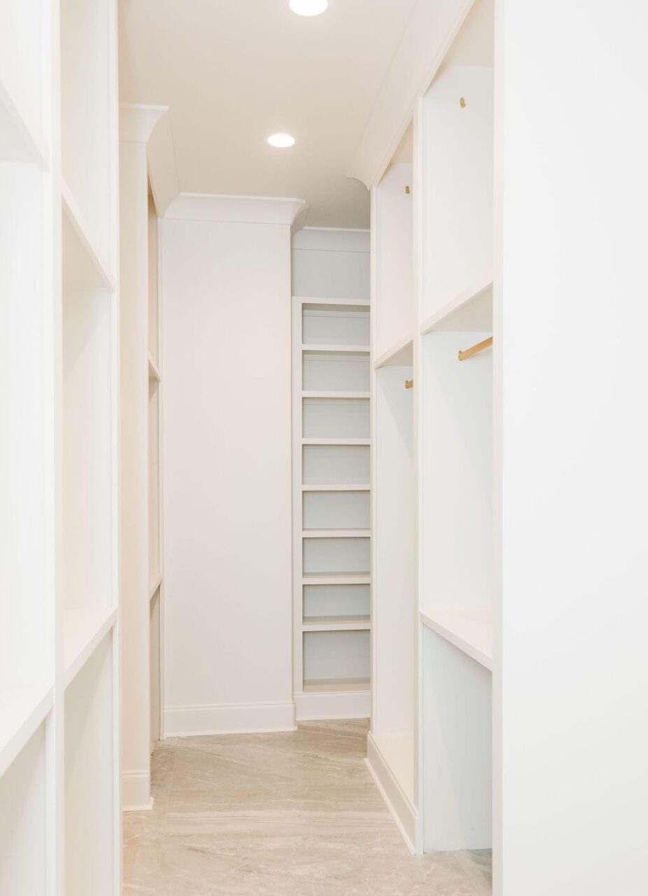 A narrow, empty walk-in closet with built-in white shelving, gold hanging rods, and recessed ceiling lights. The floor is light-colored, and there are floor-to-ceiling shelves at the end of the space.