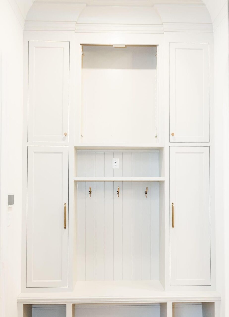 A built-in white cabinet with two tall doors on each side, three metal hooks under a shelf in the middle, an electrical outlet above the hooks, and a large open compartment at the top.