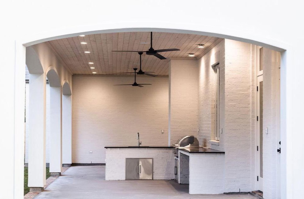 Covered outdoor kitchen with white brick walls, black countertops, a built-in grill, sink, two ceiling fans, recessed lighting, and open archways on one side. The design is modern and minimalistic.