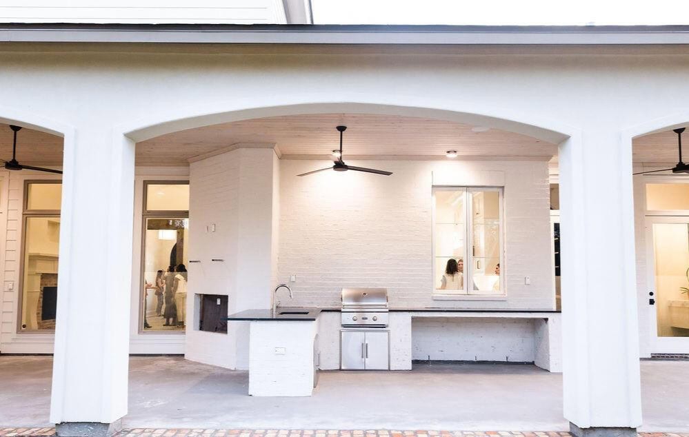 Covered outdoor patio with a built-in grill, sink, and countertop under a ceiling fan. White brick walls and large windows look into the house. The floor is unfinished concrete, and a brick border lines the patio edge.