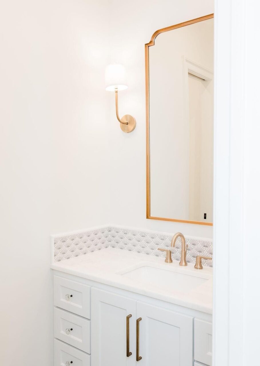 A bathroom vanity with white cabinets, a white countertop, a brass faucet, a large mirror with a brass frame, a brass wall sconce, and light patterned backsplash tiles against white walls.