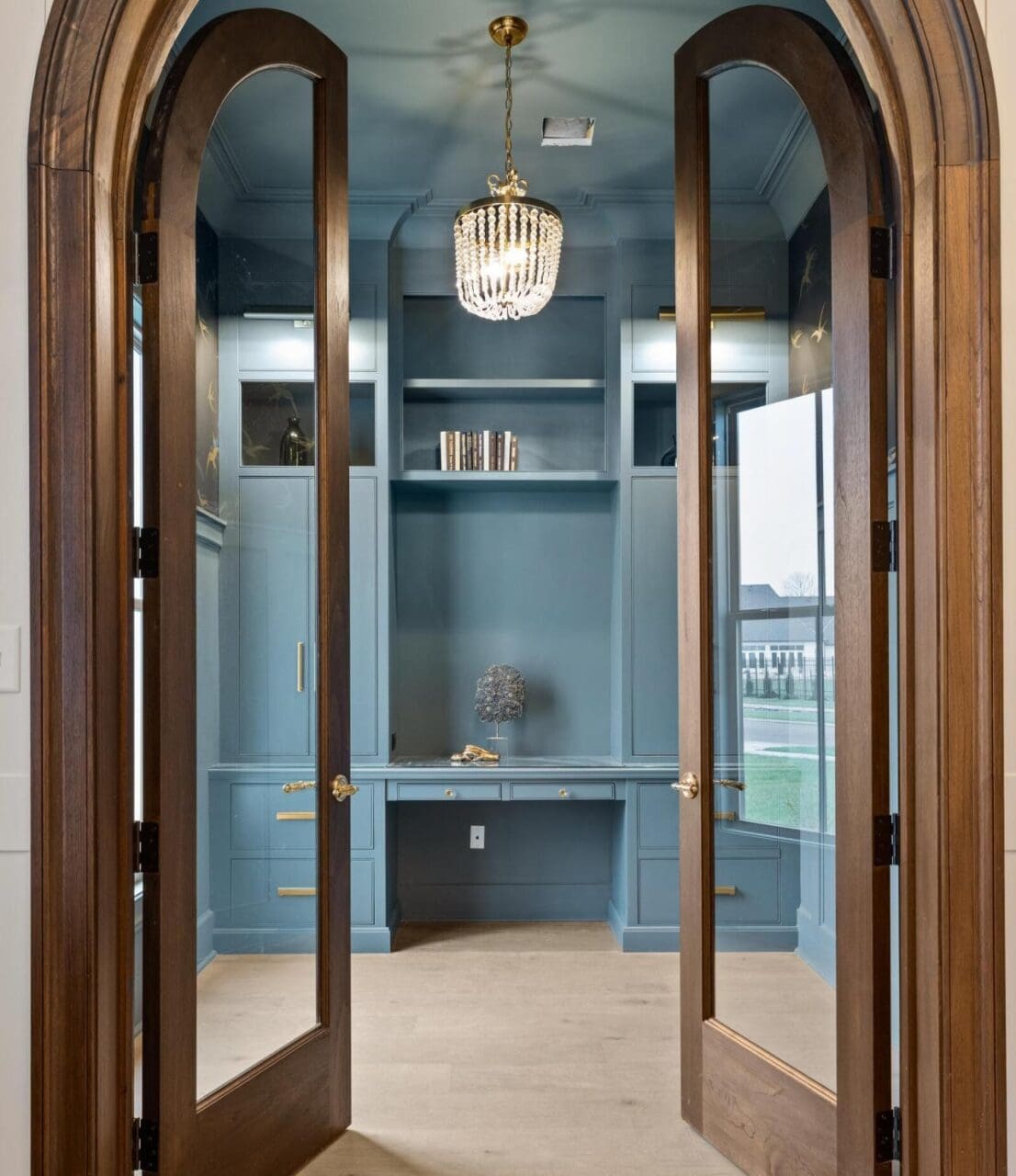 A home office with blue built-in cabinets and desk, light wood floors, and a chandelier. Two arched glass doors frame the entrance, and windows provide natural light. Shelves above the desk hold a few books.