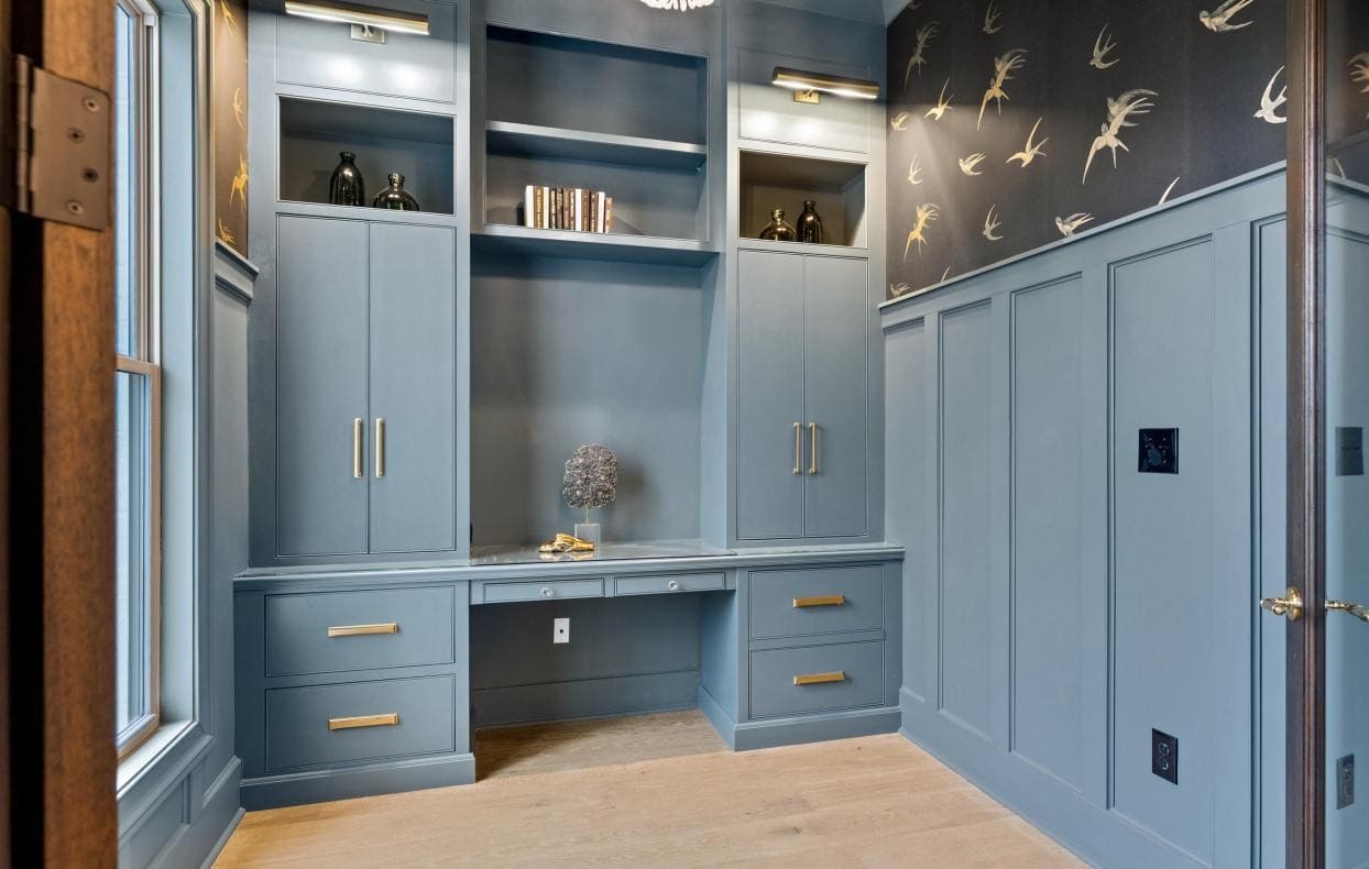 A built-in blue desk with gold handles, surrounded by cabinets and shelves, is set against a wall with bird-patterned wallpaper. The room has wood flooring and a large window on the left.