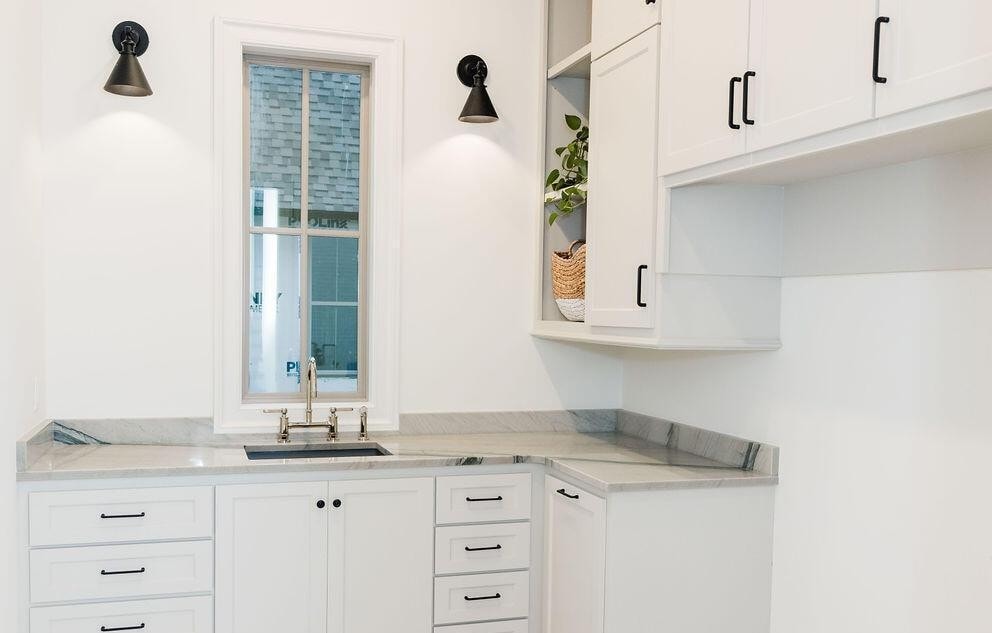 A corner kitchen with white cabinets, black handles, a marble countertop, a small window above a stainless steel sink, two black wall sconces, and a shelf with a basket and a potted plant.
