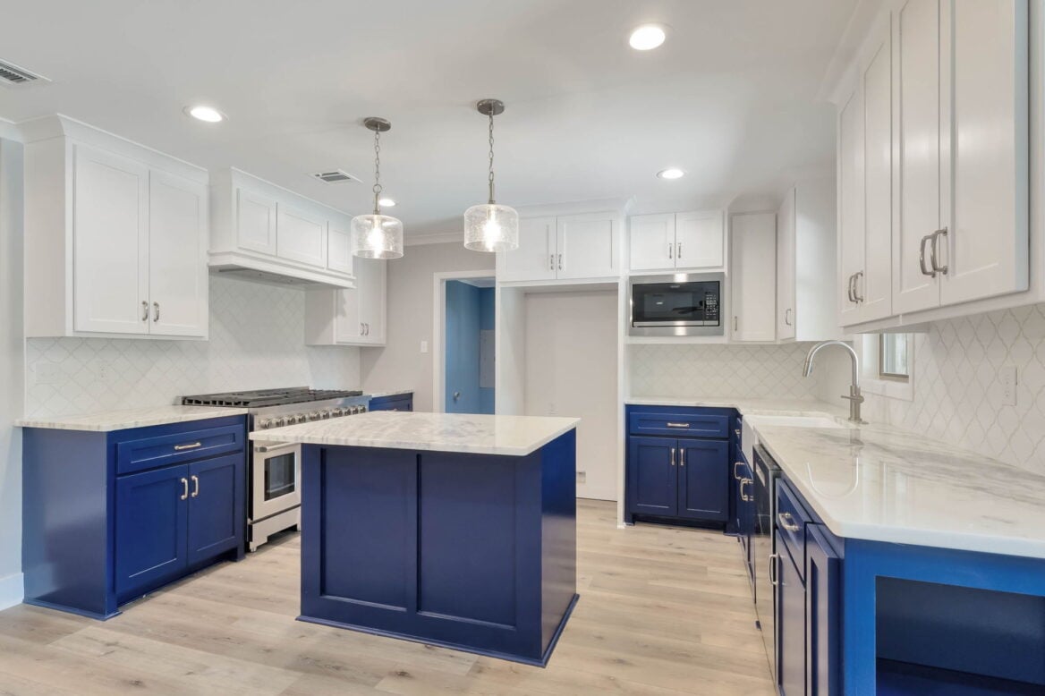 Modern kitchen with white upper cabinets, blue lower cabinets and island, white marble countertops, stainless steel appliances, pendant lights, light wood flooring, and a white tile backsplash.
