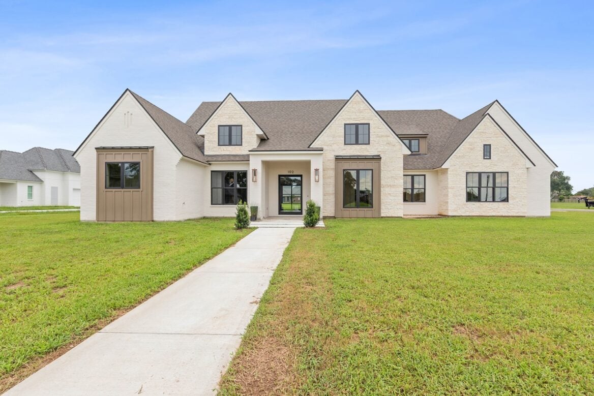 A modern, two-story house with light brick exterior, dark trim, and multiple peaked roofs. A concrete walkway leads through a large grassy yard to the front entrance, which features glass double doors.