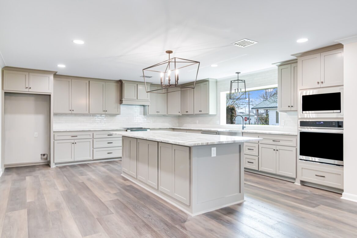 A modern, spacious kitchen with light gray cabinets, a large center island, marble countertops, stainless steel appliances, and wood flooring. Large windows provide natural light; a geometric chandelier hangs above the island.