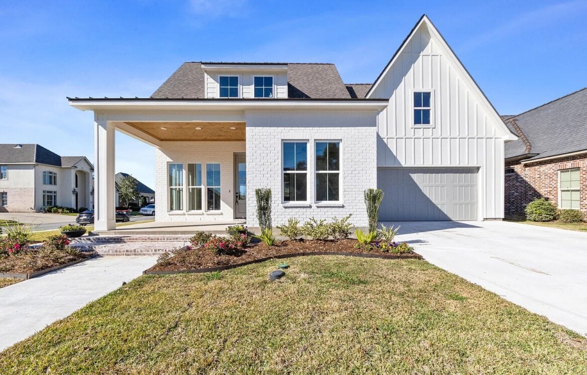 A modern two-story house with white brick and siding, a covered front porch, large windows, and an attached garage. The front yard has freshly planted shrubs and a concrete driveway under a clear blue sky.
